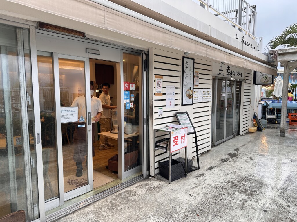 雨に濡れるウミカジテラスの外観と幸せのパンケーキの入り口。白いパラソルとテラス席が見える。
